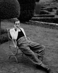 Man seated on a metal garden chair wearing a light cardigan and trousers in a formal garden.