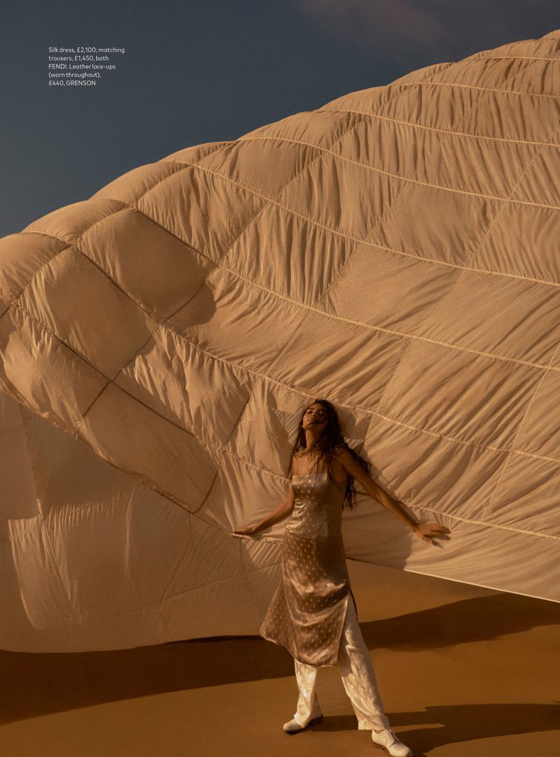 Woman in silk dress and trousers poses against a large quilt in a desert landscape under a clear sky during daytime.