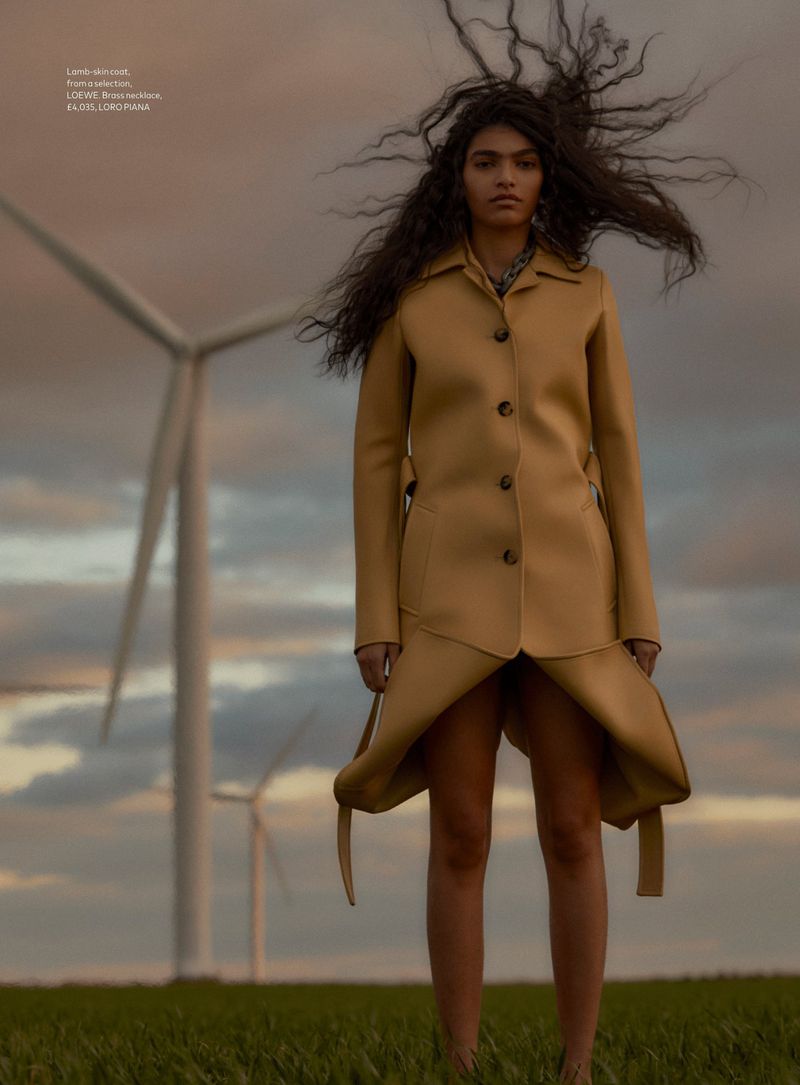 A woman in a tan coat stands in a field of green grass with wind turbines in the background, captured in a fashion-editorial style shot.