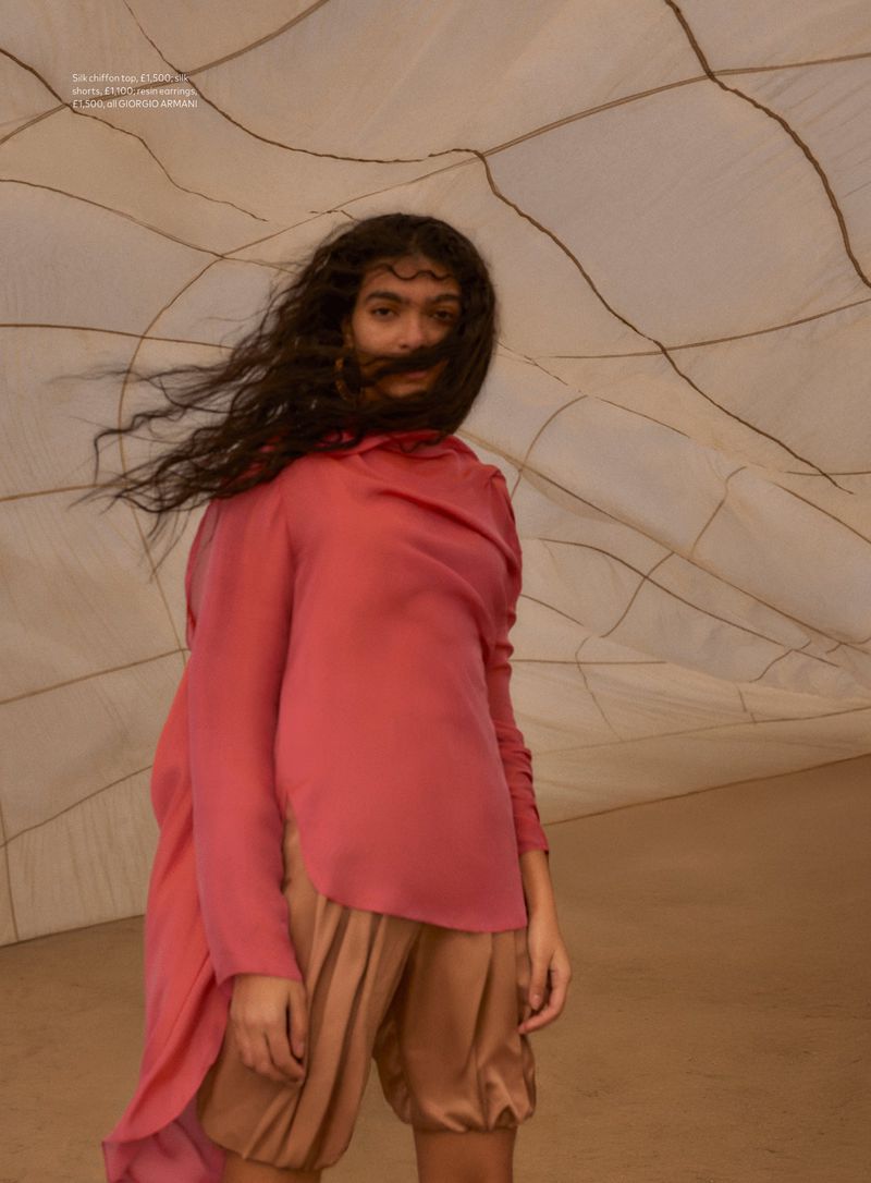 A young person with curly dark hair is modeling a long-sleeved pink top with a matching skirt and beige shorts in an indoor studio.