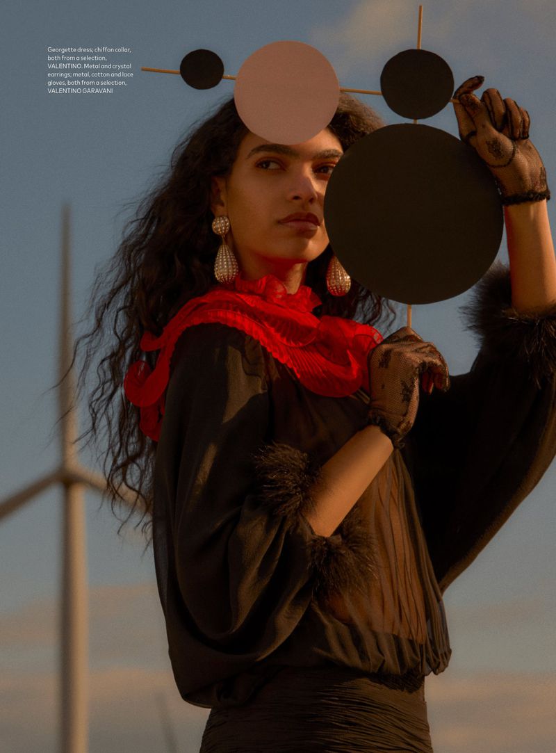 Woman in artistic fashion portrait with black dress, red scarf, statement earrings, holding a disc in outdoor setting with wind turbines.