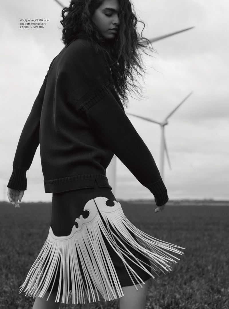 Monochrome fashion shot of a woman in a sweater and fringe skirt standing in a field with wind turbines in the background.