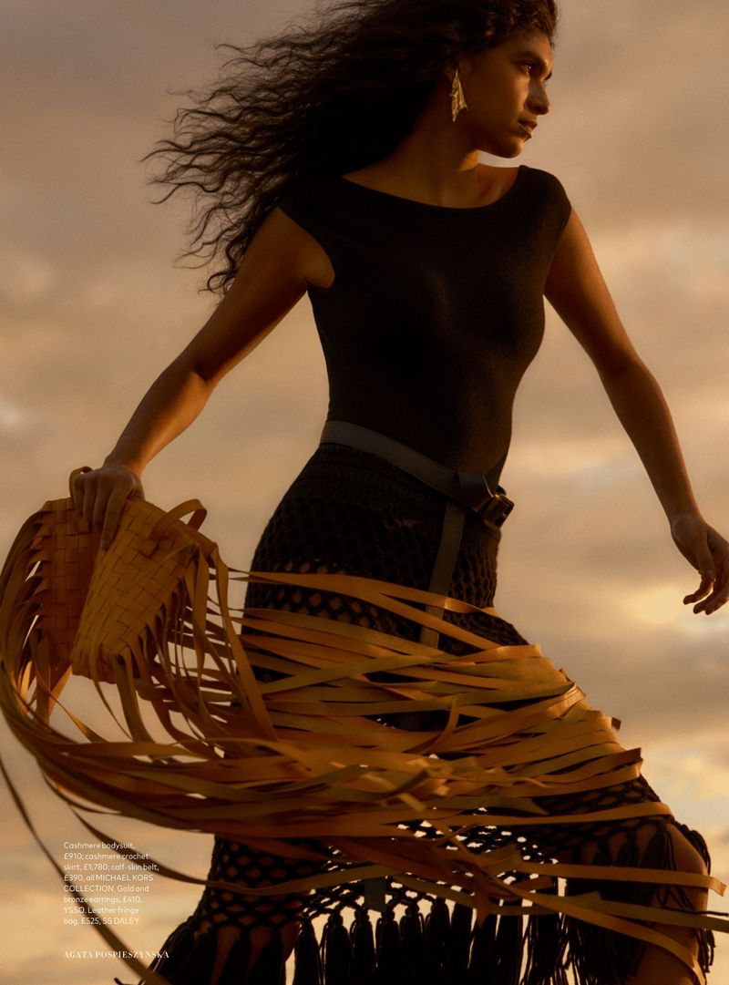 Fashion editorial shot of an adult woman wearing a black top, skirt with tan fringe, and carrying a tan woven bag in an outdoor setting.