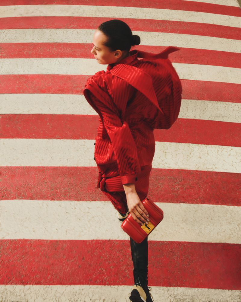 Fashionable woman in red outfit crossing a red and white striped crosswalk with a red clutch, turning her head back to look behind her.