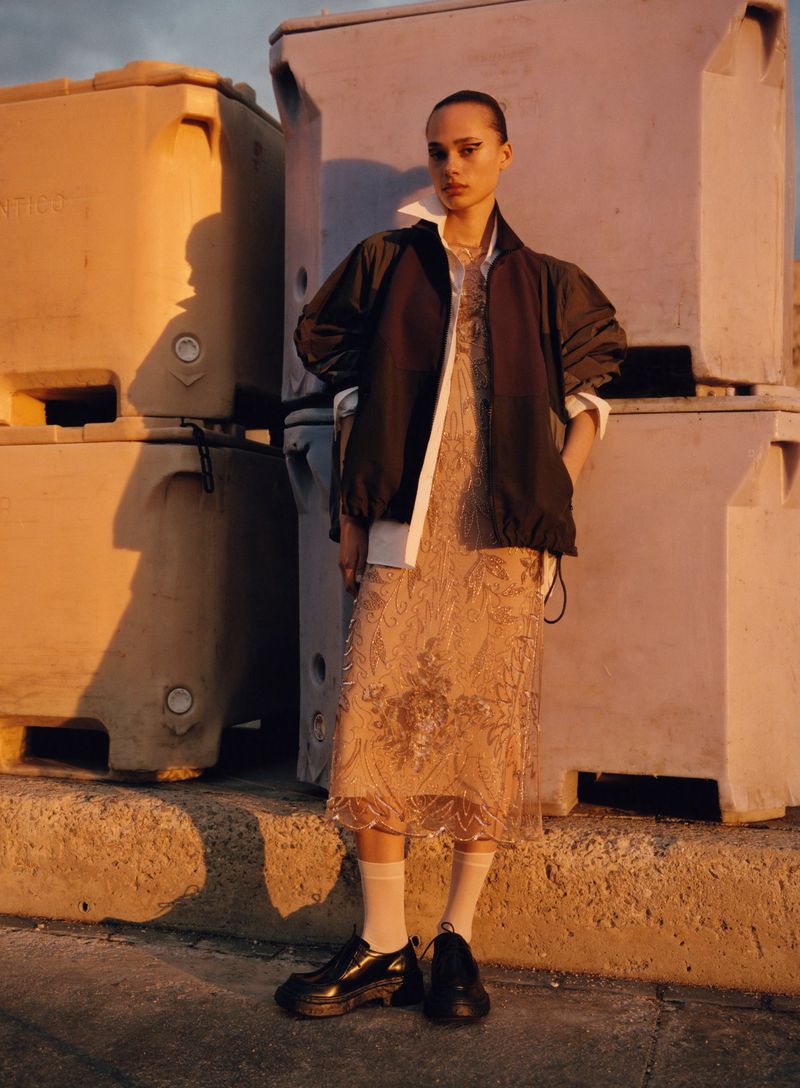A woman in a fashionable brown jacket and patterned dress stands outdoors with her hands in her pockets, in soft, warm lighting.