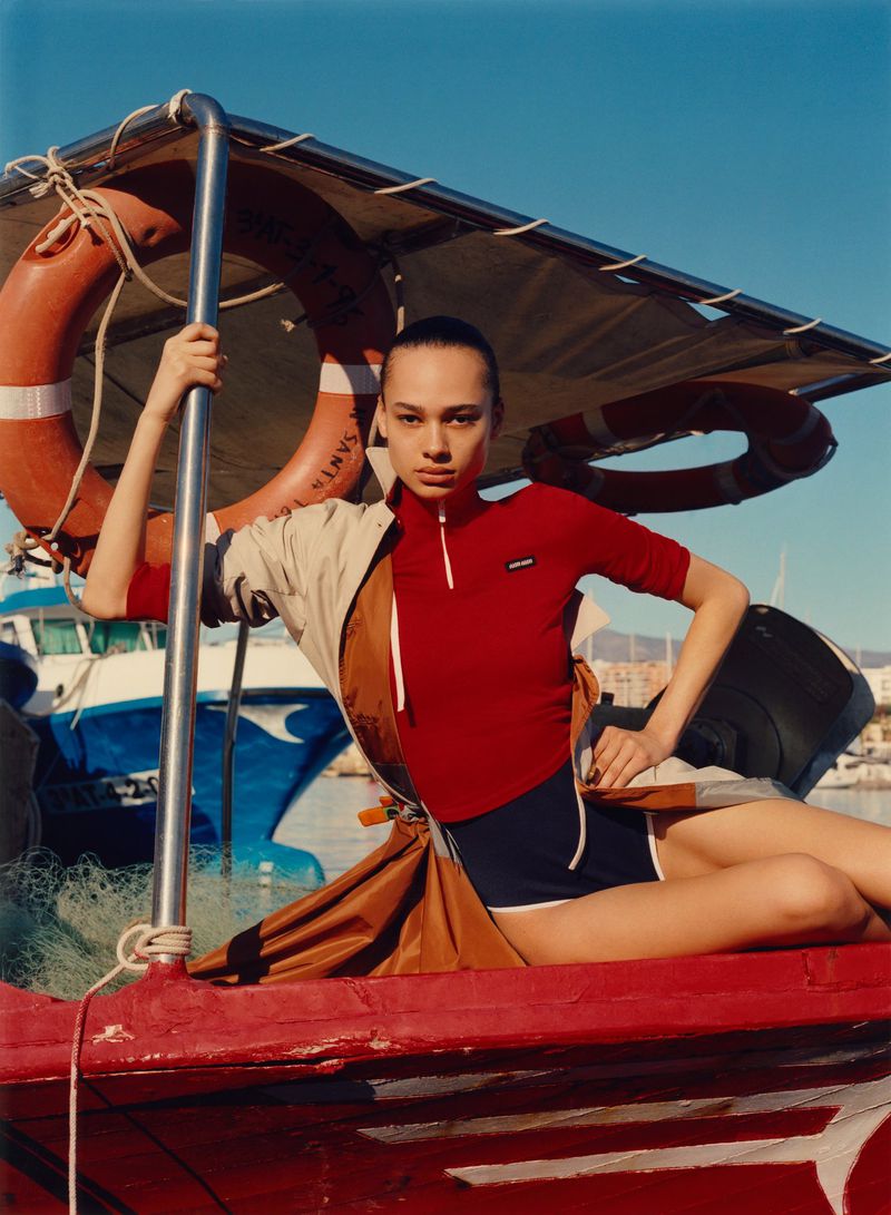 A model on a red boat wears a sporty red top and navy shorts with life preserver and blue boat visible in the background on a sunny day.
