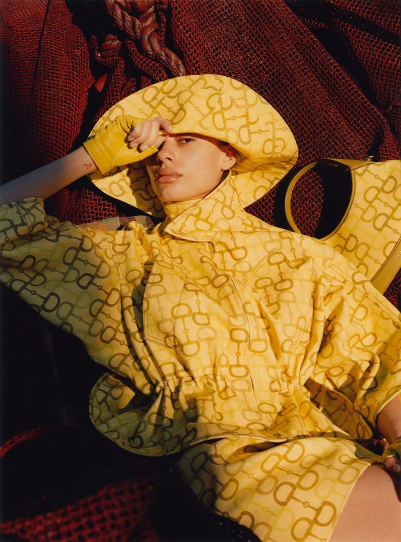 Woman in yellow patterned matching set with hat and purse lays in a red net with hand shielding her eyes in a fashion editorial.