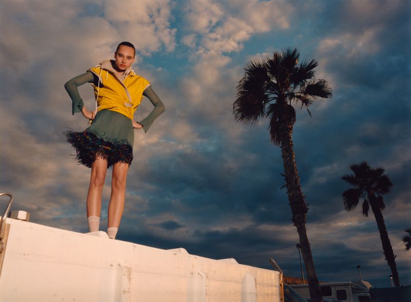 Fashionable young woman stands on a rooftop at dusk with palm trees against a cloudy sky, wearing a yellow vest and green fringe skirt.