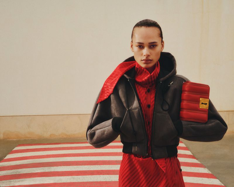 Fashion portrait of a woman in a grey jacket with a red purse and dress, posing against a minimalist wall and red striped floor.