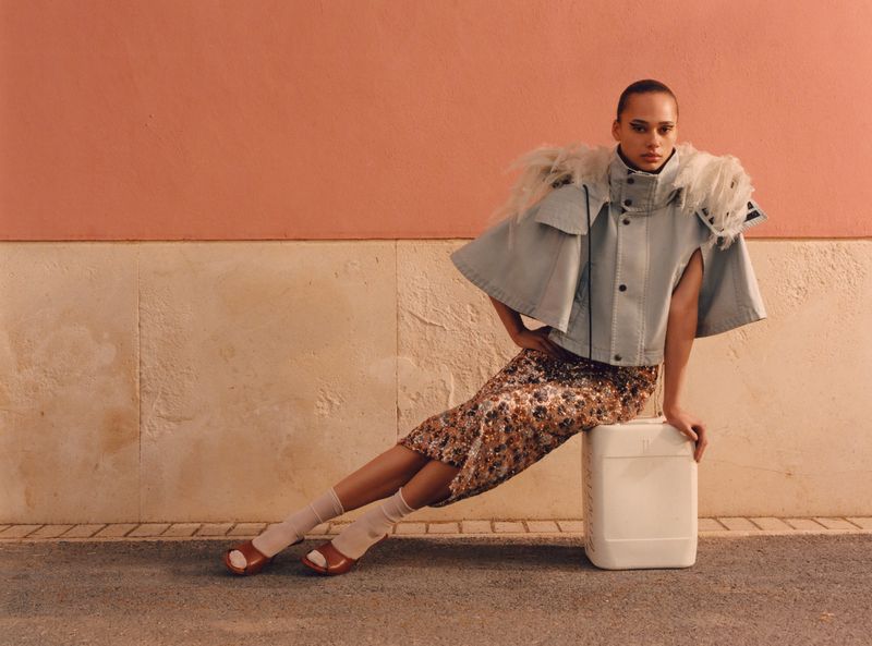 An adult female poses while sitting on a container wearing a midi skirt and denim jacket, in front of a peach and beige wall.