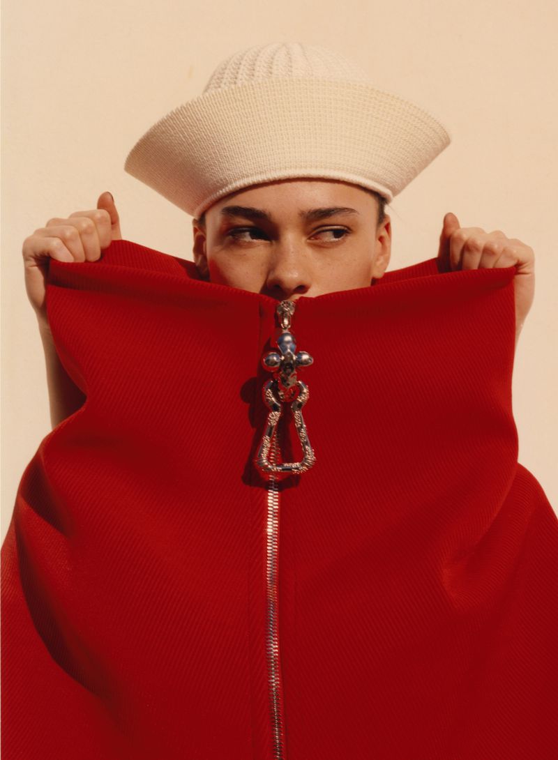 Portrait of model wearing a white cap and red garment with decorative zipper pull, glancing off to the left against a cream background.