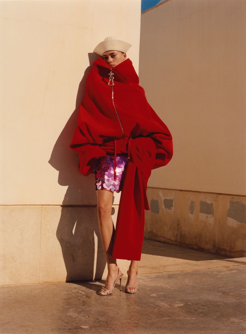 Fashion photo of a non-binary person in a red oversized coat with a gold zipper, sequin skirt, strappy heels, and a hat against a wall.