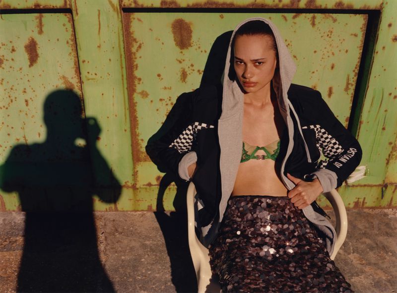 Young woman with sequin skirt and jacket sits on chair in front of green metal background with shadow of person visible.
