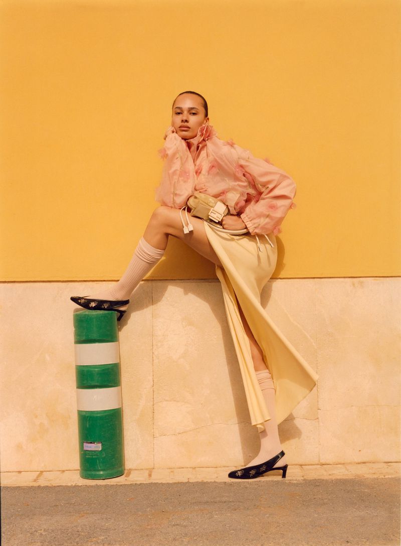 Adult woman in pink jacket and cream skirt poses with one leg on a green and white bollard against a yellow wall on a city street.
