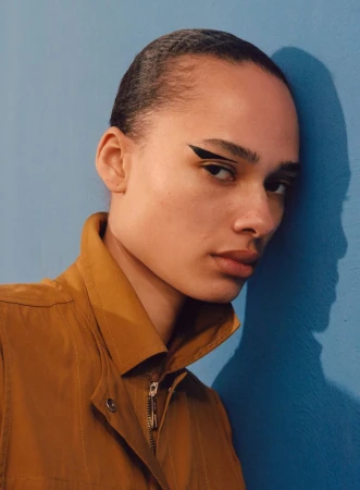 Model wearing a brown jacket with a prominent collar leaning against a blue wall with graphic eyeliner.
