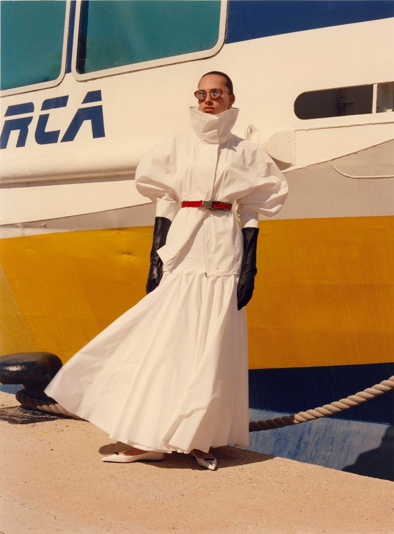 Model wearing a white structured coat and black gloves standing in front of a large painted ship hull