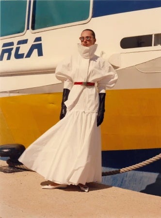 Model wearing a white structured coat and black gloves standing in front of a large painted ship hull