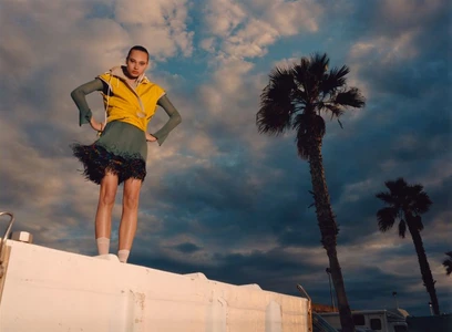 Model wearing a yellow vest and green skirt standing on a ledge against a sunset sky with palm trees.