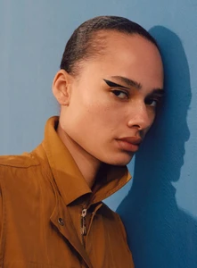 Model wearing a brown jacket with a prominent collar leaning against a blue wall with graphic eyeliner.