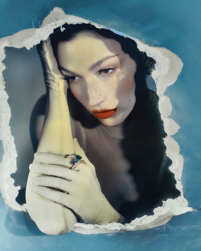Portrait of a woman with fair skin, red lips, and dark hair, framed by a torn paper effect with blue backdrop and wearing a blue gemstone ring.