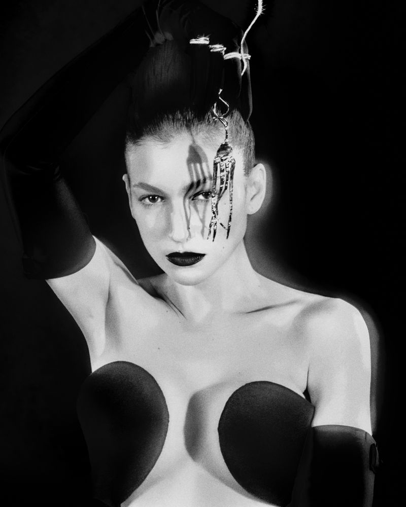 Monochrome portrait of an adult female with dramatic makeup and a fork prop over her face, set against a dark background, studio shot.