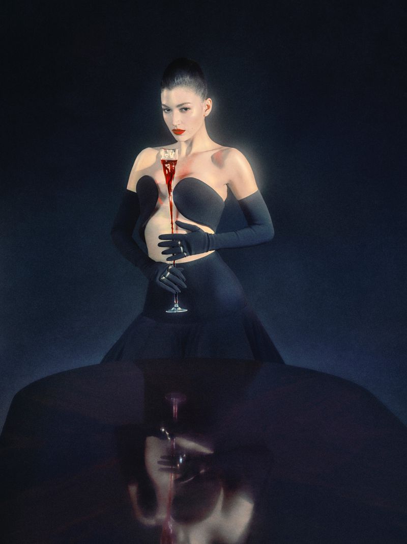 Elegant woman in black dress and gloves, holding glass with red liquid, posing in dark studio with reflection; beauty and fashion.