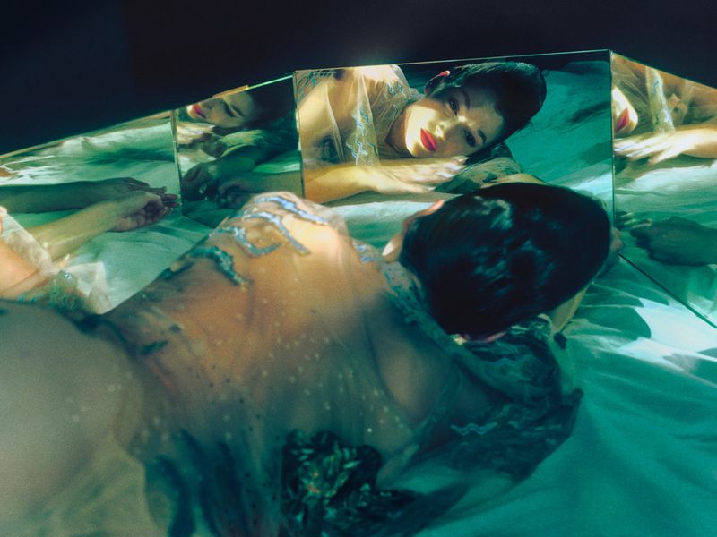 Woman lying on bed, reflected in multiple mirrors, wearing a sheer top with silver details, red lipstick, and short dark hair.
