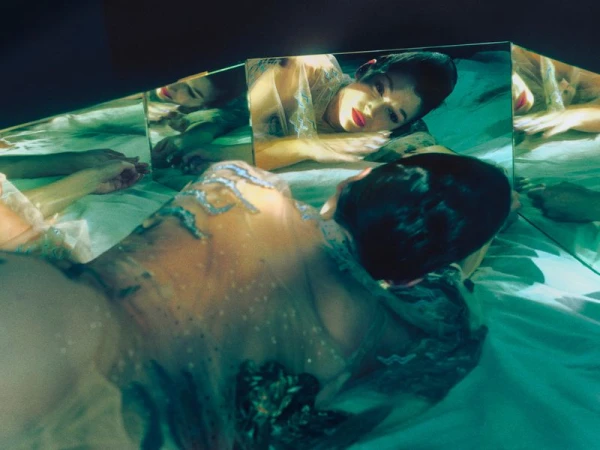 Woman lying on bed, reflected in multiple mirrors, wearing a sheer top with silver details, red lipstick, and short dark hair.
