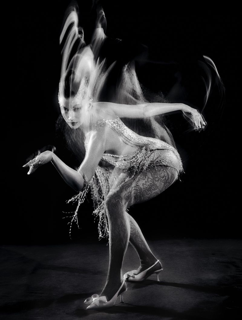 Monochrome long-exposure portrait of a woman in fringed flapper dress, bending at the knee with arms outstretched on black background.