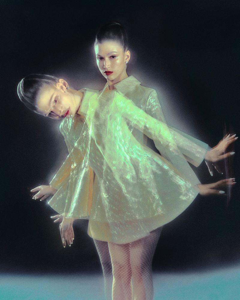 Fashion photography of an adult female with a semi-transparent trench coat and multiple exposure effect in front of a dark background.