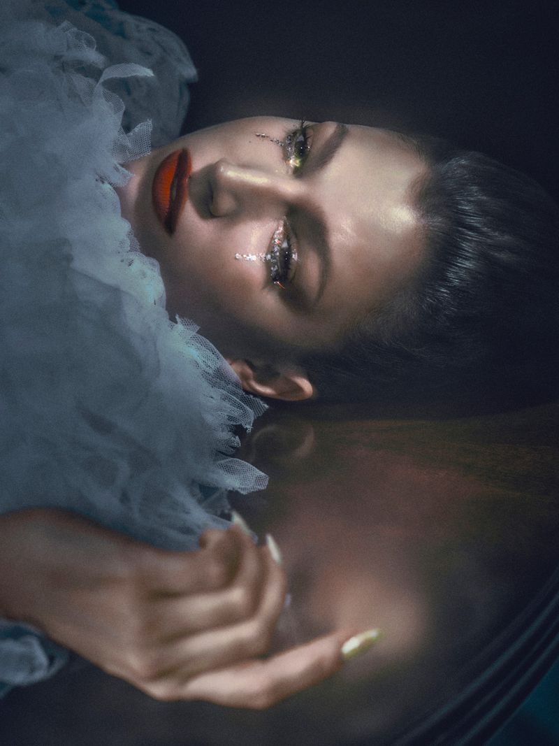 Dramatic portrait of a person with glitter makeup lying in water. The artistic lighting and the reflection enhance the surreal quality.