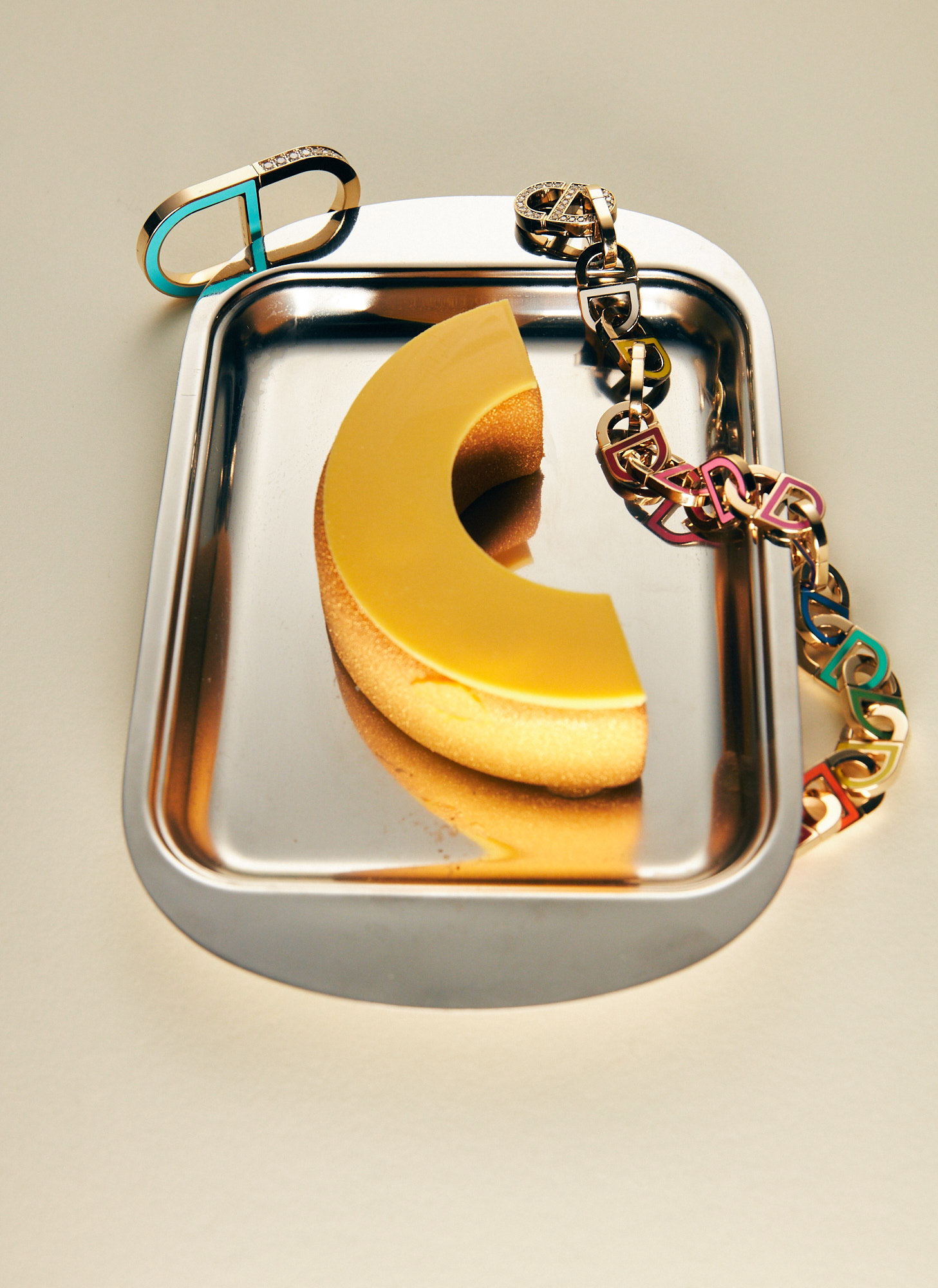 Still life shot on beige backdrop features dessert slice on a silver tray, plus bracelet and chunky chain jewelry.