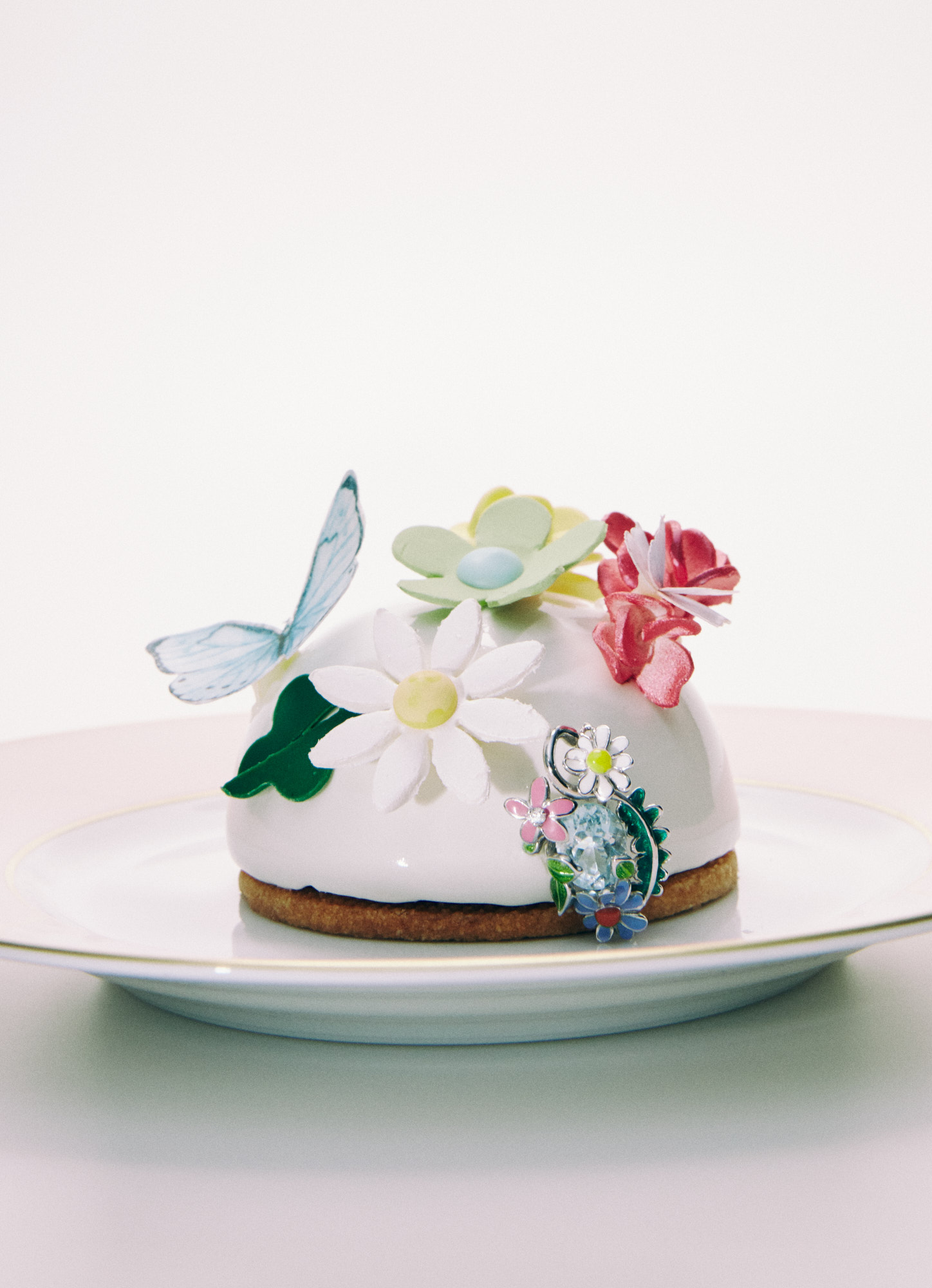 A white domed dessert is adorned with colorful flowers and a butterfly, resting on a white plate against a bright white backdrop.
