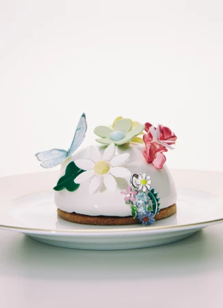 A white domed dessert is adorned with colorful flowers and a butterfly, resting on a white plate against a bright white backdrop.