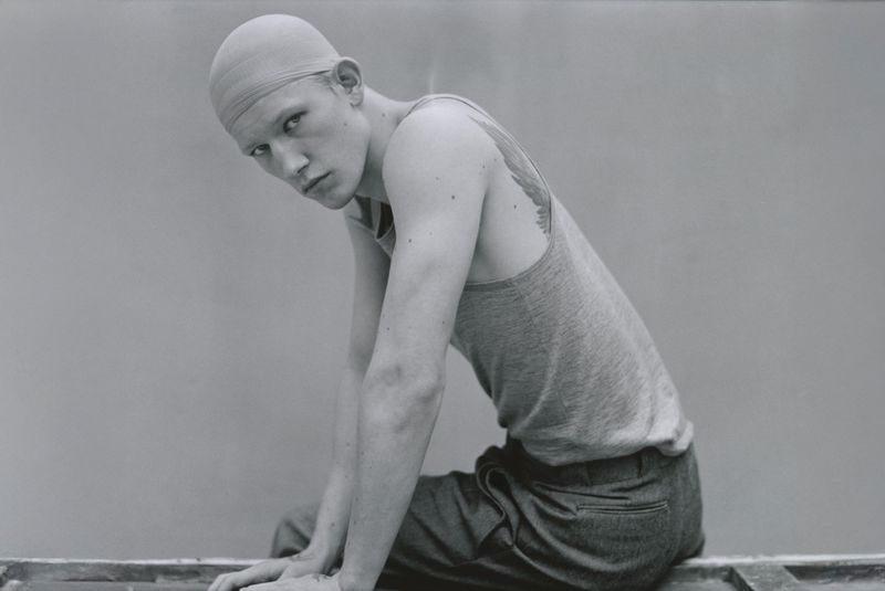 Monochrome portrait of a young adult male model in a cap and casual tank top, sitting indoors with a neutral backdrop.