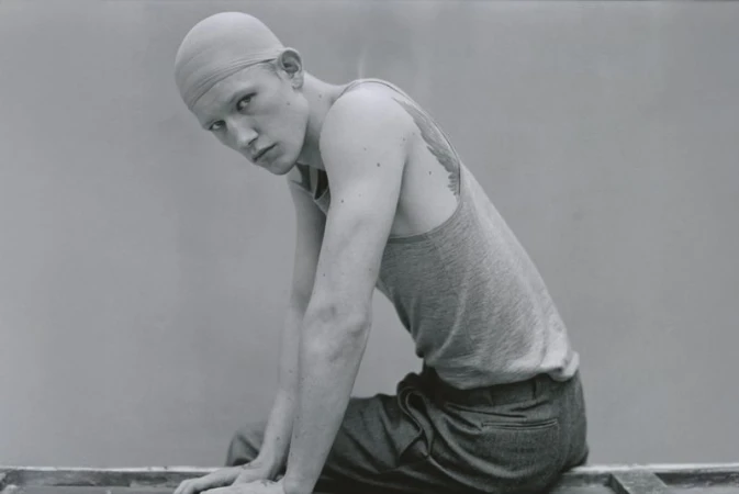 Monochrome portrait of a young adult male model in a cap and casual tank top, sitting indoors with a neutral backdrop.