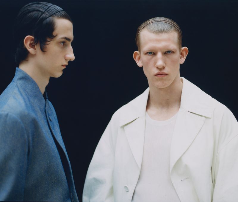 Two men pose indoors against a dark backdrop. One man looks directly at the camera wearing a white jacket and the other wears denim.