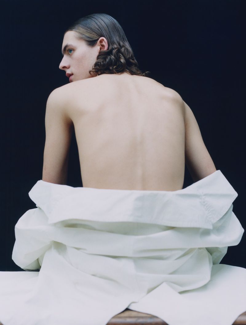 Back view of an adult male with dark wavy hair, no shirt, and a white jacket around his waist.  He's seated against a dark studio backdrop.