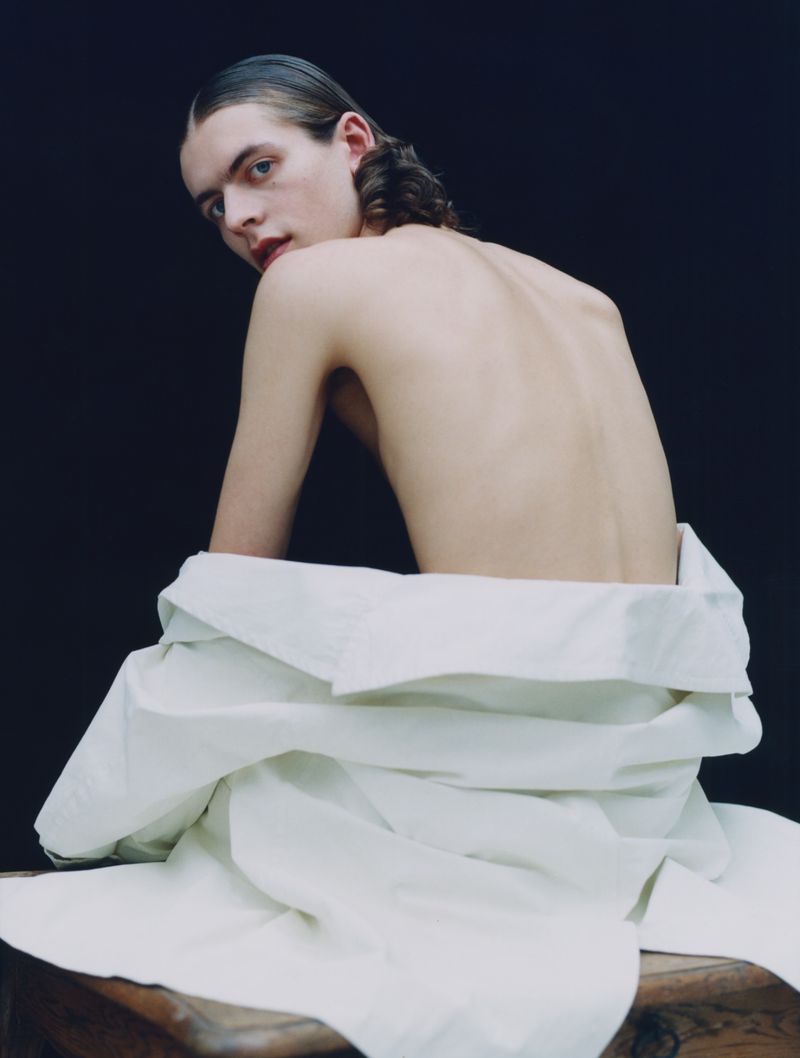 Portrait of a topless young adult male with slicked-back hair sitting on a wooden object, wearing a white shirt and posing against a dark background.