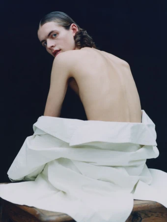 Portrait of a topless young adult male with slicked-back hair sitting on a wooden object, wearing a white shirt and posing against a dark background.