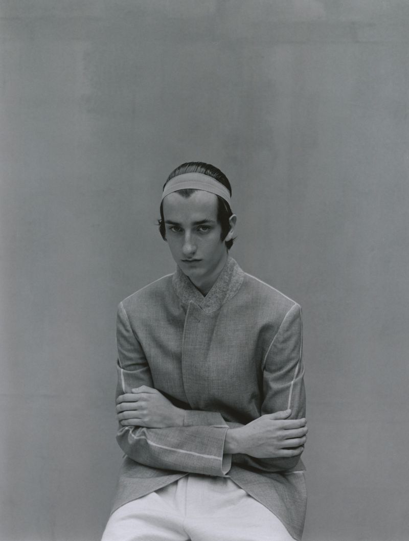 A portrait of a young adult male wearing a gray jacket, white pants, and a headband, with his arms crossed in a neutral indoor setting.