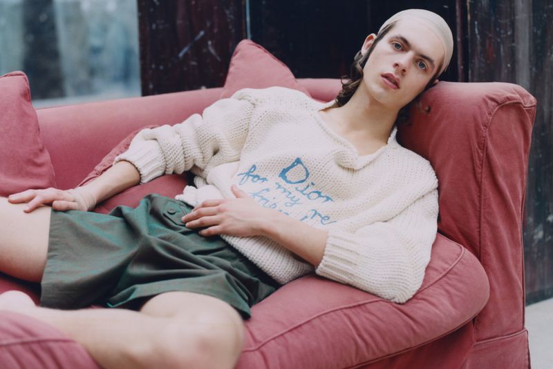 Young adult male reclines on a pink couch wearing a cream-colored sweater with green shorts, indoor setting with a dark marble-like background.