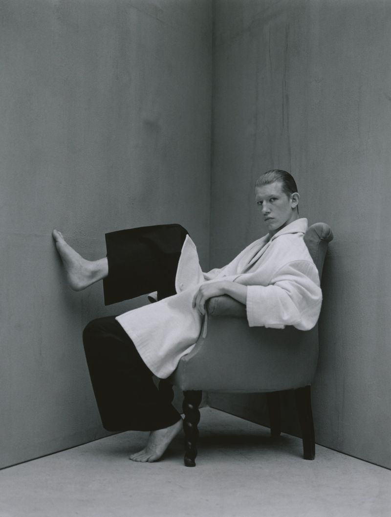 A young adult male sits in a wingback chair in a grey room wearing a white bathrobe, looking at the camera with a neutral expression.
