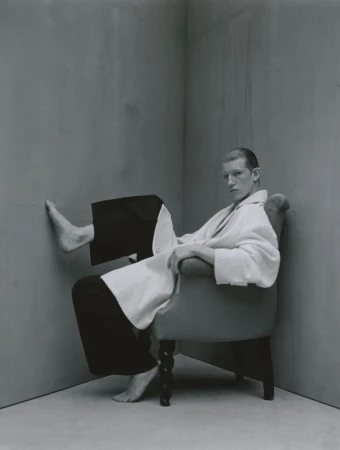 A young adult male sits in a wingback chair in a grey room wearing a white bathrobe, looking at the camera with a neutral expression.