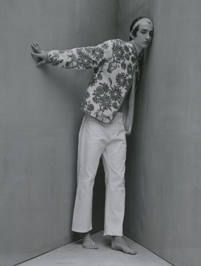 Young man in printed jacket and white trousers poses in a corner, captured in a monochromatic, full shot highlighting fashion and form.