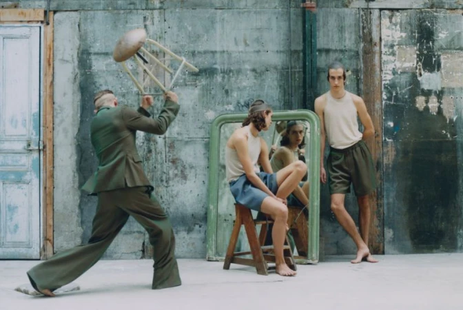 Three men in an indoor setting, one with a chair overhead, another seated by a mirror, and the third standing next to a wall.