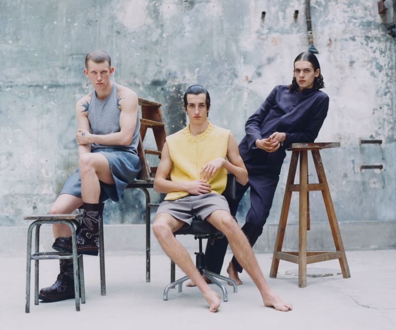 Three young adult men pose in a photo studio against a grey textured wall. One man sits in an office chair, one sits on a stool and the other leans on a tall stool.