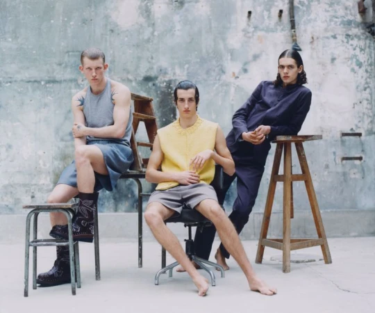 Three young adult men pose in a photo studio against a grey textured wall. One man sits in an office chair, one sits on a stool and the other leans on a tall stool.