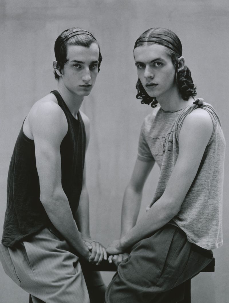 Two young male models sitting together wearing tank tops against a grey studio backdrop for Lampoon Magazine project Between us three.