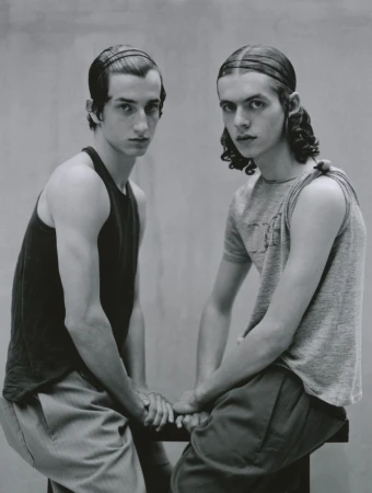 Two young male models sitting together wearing tank tops against a grey studio backdrop for Lampoon Magazine project Between us three.